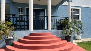 A building with a blue exterior, a black door, and a set of red steps leading to a covered porch with white columns. - Tranquil 2-bedroom apartment in Beautiful Barbados (Saint Philip)