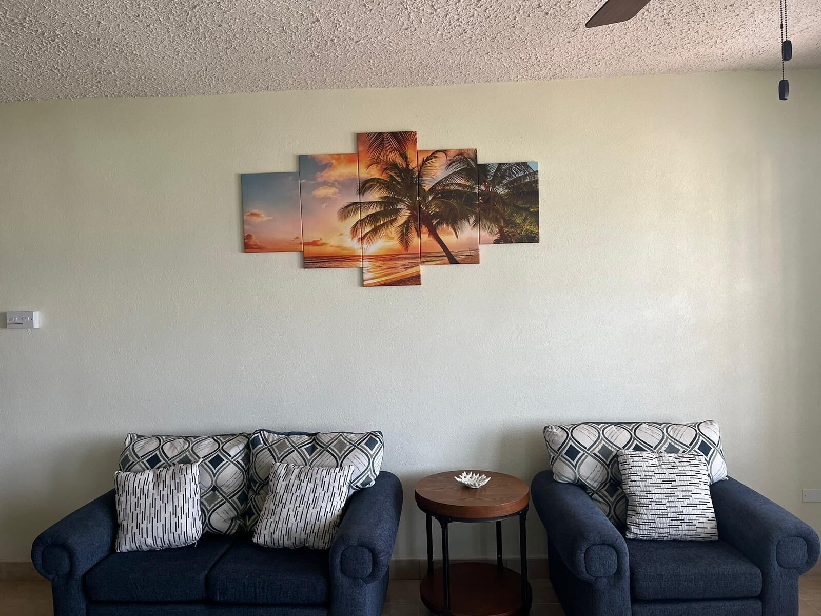 Un salon avec deux fauteuils bleus, des coussins à motifs et un tableau représentant un coucher de soleil, accroché au mur.
