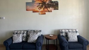 A living room with two blue armchairs, patterned cushions, and a wall-mounted sunset painting. - Tranquil 2-bedroom apartment in Beautiful Barbados (Saint Philip)