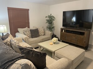 Un salon avec un canapé beige, des coussins à motifs, une table basse sur laquelle est posée une bougie, un téléviseur sur un meuble en bois et une plante en pot.