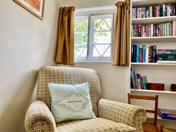 A room with a patterned armchair, a bookshelf with various books, a window with curtains, and a framed picture on the wall.