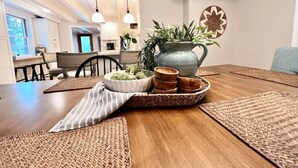 Un comedor con una mesa de madera dispuesta para una comida, un florero con flores y vista a la cocina al fondo.