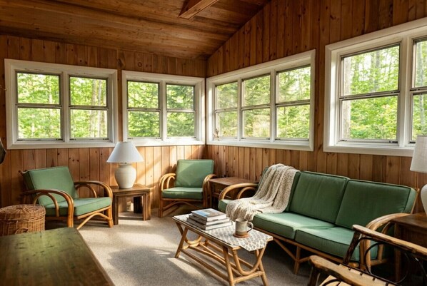 A cozy cabin interior with wooden walls, large windows, a green sofa, wicker chairs, and a small table with books and a mug.