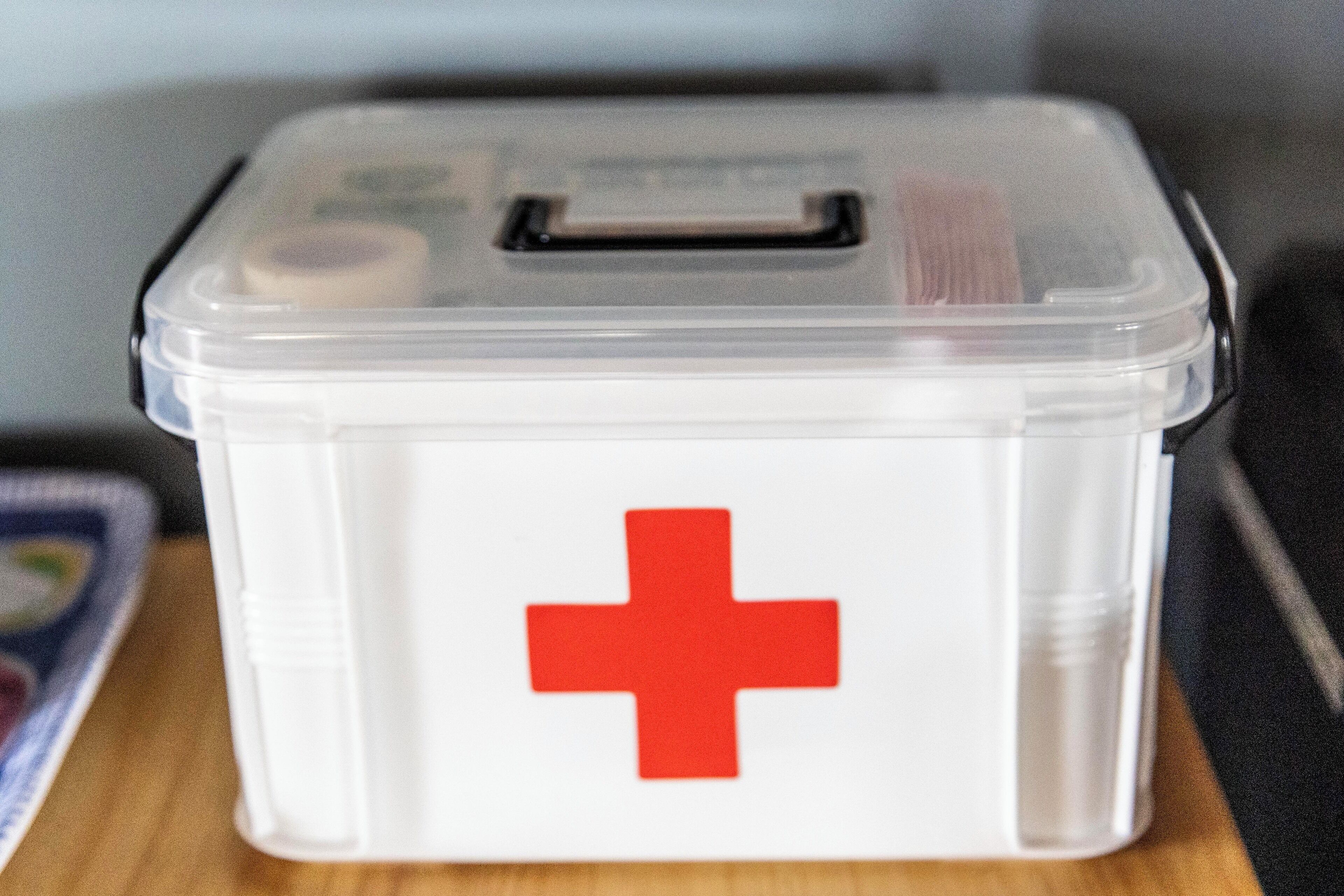 A clear plastic storage box with a red cross symbol on it, placed on a wooden surface.