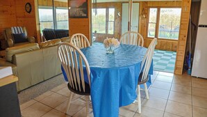 A wooden cabin interior with a dining table covered in a blue tablecloth, surrounded by white chairs, and a sofa with a striped cushion. - Catch on the Canal - Waterfront Getaway Near Casino, Dining & Entertainment (Standish)