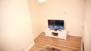 A room with a flat-screen TV on a white stand, a small shelf with red items, and a wooden floor. - Lovely 1-bedroom condo in Greater London (London)