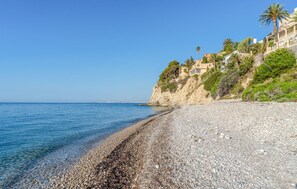 A pebble beach with a clear blue sea, a rocky cliff, and buildings with balconies overlooking the beach.