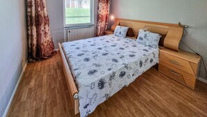 Een slaapkamer met een houten bed, een nachtkastje, een raam met gordijnen en een radiator.