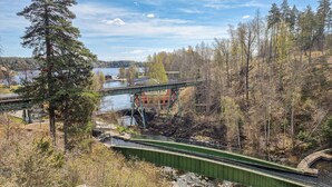 Een groene voetgangersbrug over een rivier, met een bebost gebied en gebouwen op de achtergrond.