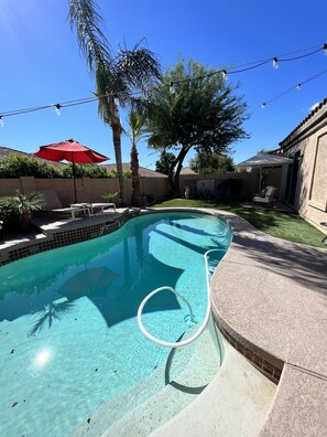 Une piscine à l’eau d’un bleu limpide, entourée d’une terrasse agrémentée d’un parasol rouge, d’une chaise et d’une petite table.