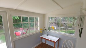 Quarto com grandes janelas, piso de madeira, pequena mesa com um vaso de flores e uma cadeira branca.