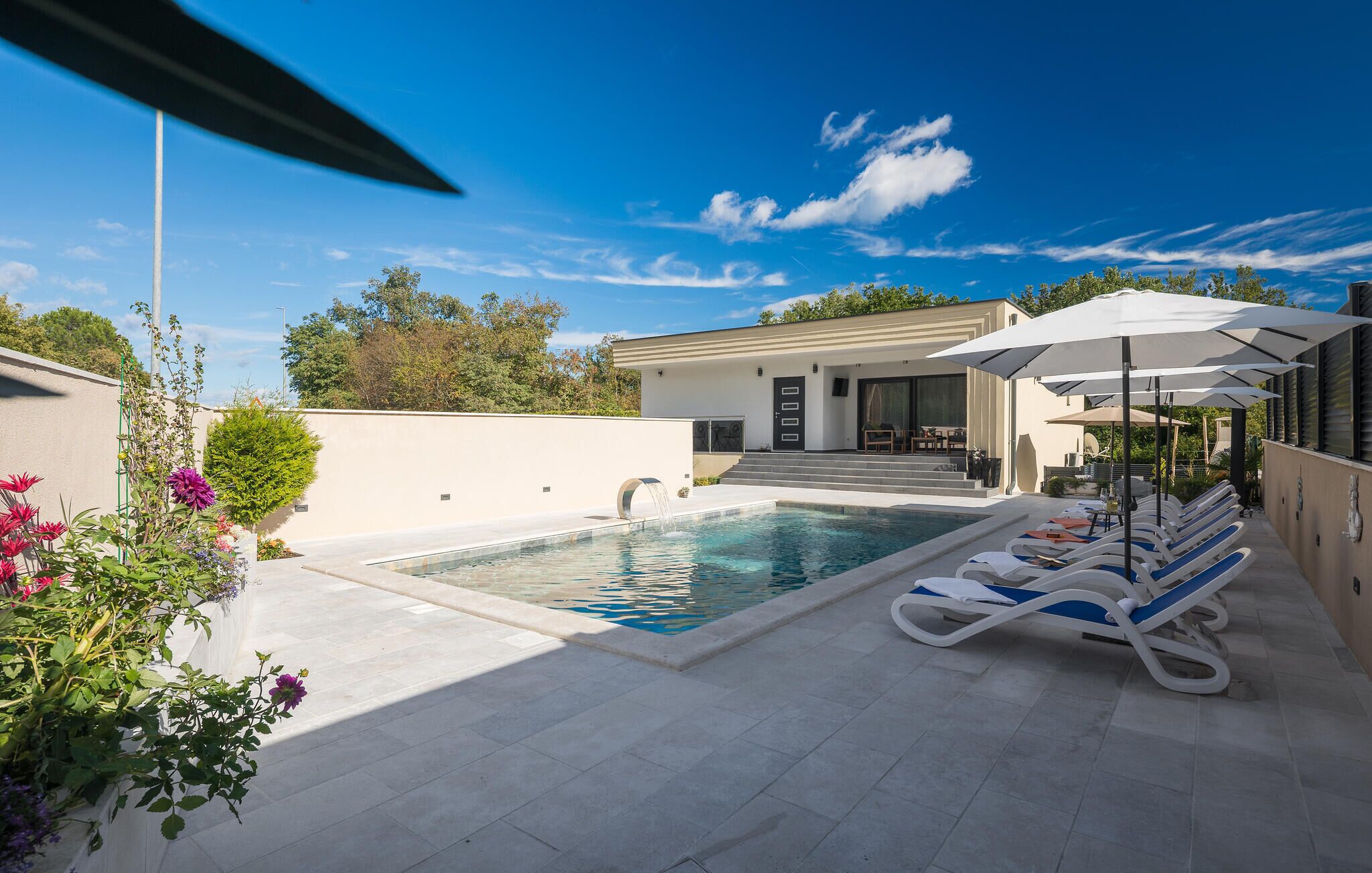 Een modern buitenzwembad met ligbedden en parasols, een helderblauwe lucht en een gebouw met minimalistisch design.
