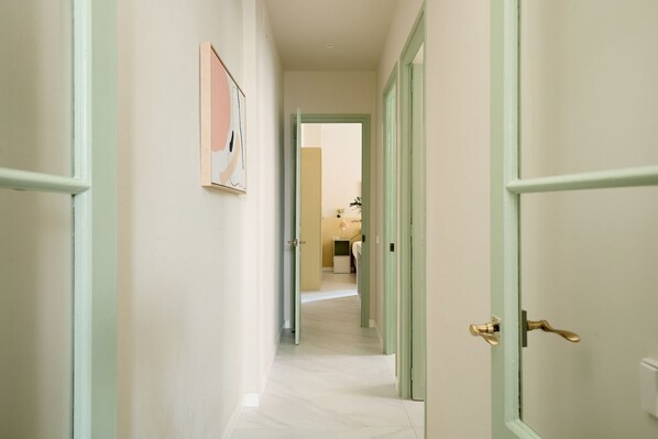 A hallway with light green doors, a framed artwork on the wall, and a view into a room with a bed and a nightstand.