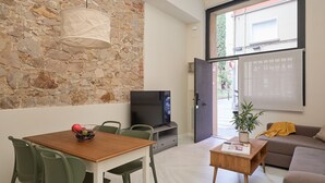 A modern living room with a stone wall, a dining area with a table and chairs, a television, a sofa, and a window with a view of greenery.