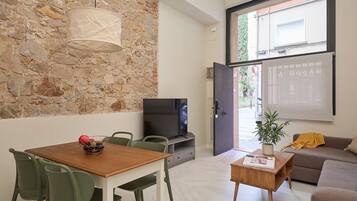 A modern living room with a stone wall, a dining area with a table and chairs, a television, a sofa, and a window with a view of greenery.