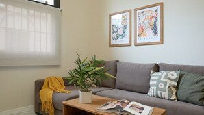 Een woonkamer met een grijze bank, een houten salontafel met een tijdschrift, een gele plaid, een potplant en twee kunstwerken in lijsten aan de muur.