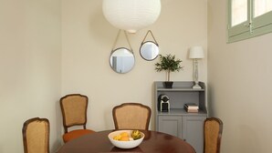 A dining area with a round table, four wicker chairs, a bowl of fruit, two wall-mounted mirrors, a hanging white lantern, a potted plant, a lamp, and a gray cabinet with books and a clock. - Casa Cosi - El Farro 4 (Barcelona)
