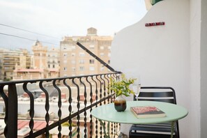 Een balkon met een metalen hekwerk, een kleine tafel met een bloevas en een boek, en een uitzicht op gebouwen op de achtergrond.