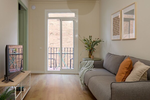 Een woonkamer met een grijze bank, een klein bijzettafeltje met een plant, een televisie op een standaard en een schuifdeur die leidt naar een buitenbalkon.