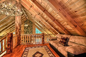 A cozy loft-style room with a wooden ceiling, a stone wall, a wooden railing, a cushioned sofa, and a patterned rug.