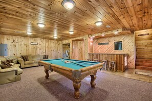 A wooden interior with a pool table, sofas, and a bar area.
