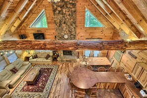 Interior acogedor de una cabaña con chimenea de piedra, paredes de madera, comedor y un ventanal grande.