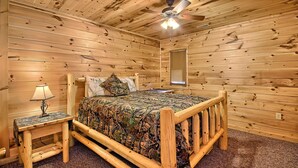Un dormitorio de cabaña de madera con una cama, una mesita de noche, una lámpara, un ventilador de techo y una ventana con persianas.