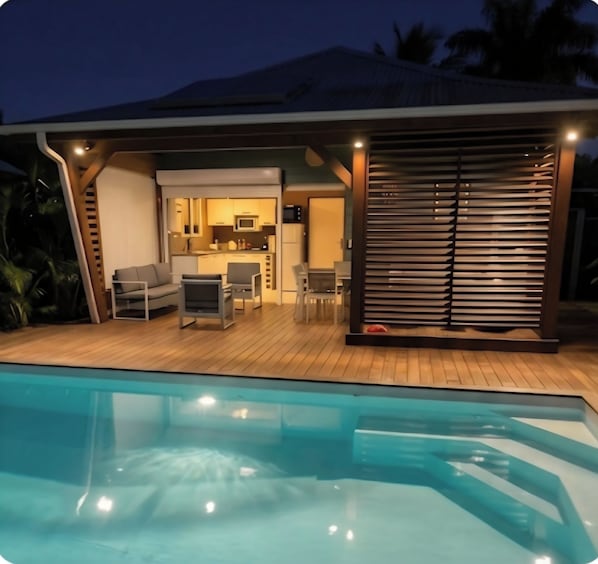 Un espacio junto a la piscina con piscina, cocina al aire libre techada y una zona de estar con sillas y mesa.