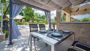 Un espace de restauration en plein air avec une table en verre, des chaises et une vue sur une piscine et la verdure.