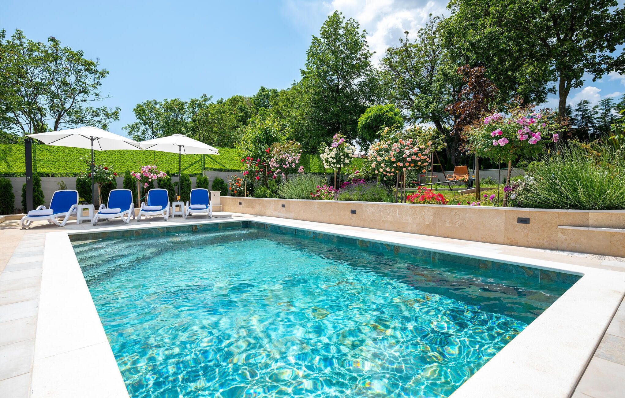 A rectangular swimming pool with clear blue water, surrounded by a tiled deck. There are three lounge chairs with blue cushions and white armrests, and two white umbrellas providing shade. The pool is set against a backdrop of lush greenery and flowering plants.