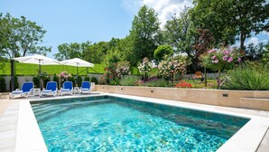 Une piscine rectangulaire à l’eau d’un bleu limpide, entourée d’une terrasse carrelée. On y trouve trois chaises longues dotées de coussins bleus et d’accoudoirs blancs, ainsi que deux parasols blancs qui offrent de l’ombre. La piscine est mise en valeur par un décor luxuriant de verdure et de plantes en fleurs.