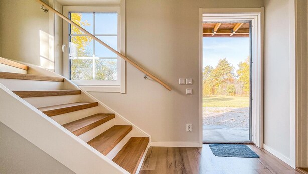 Una escalera de madera con barandilla, que lleva a una puerta con vistas a árboles y un sendero de grava.