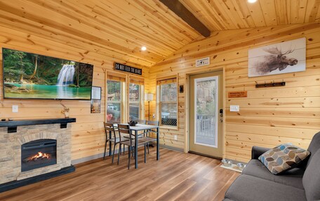 A wooden cabin interior with a fireplace, dining area, and a large window overlooking a forest.