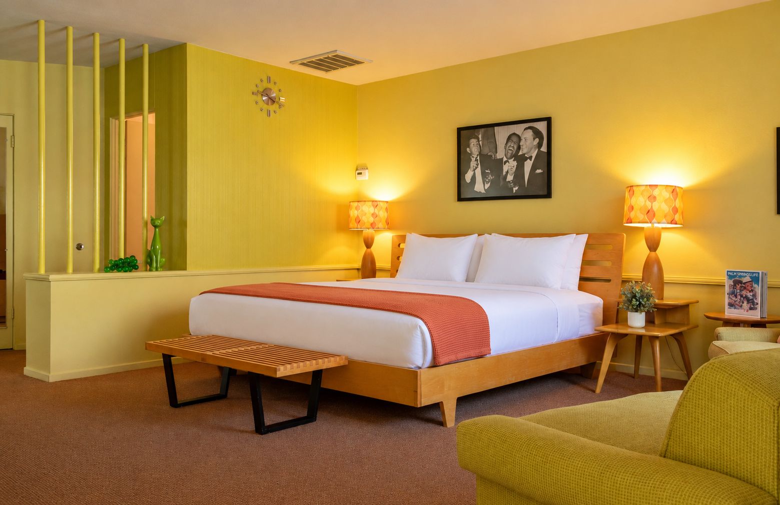 A hotel room with a bed, bedside lamps, a bench, a small table with a plant, and a picture on the wall.