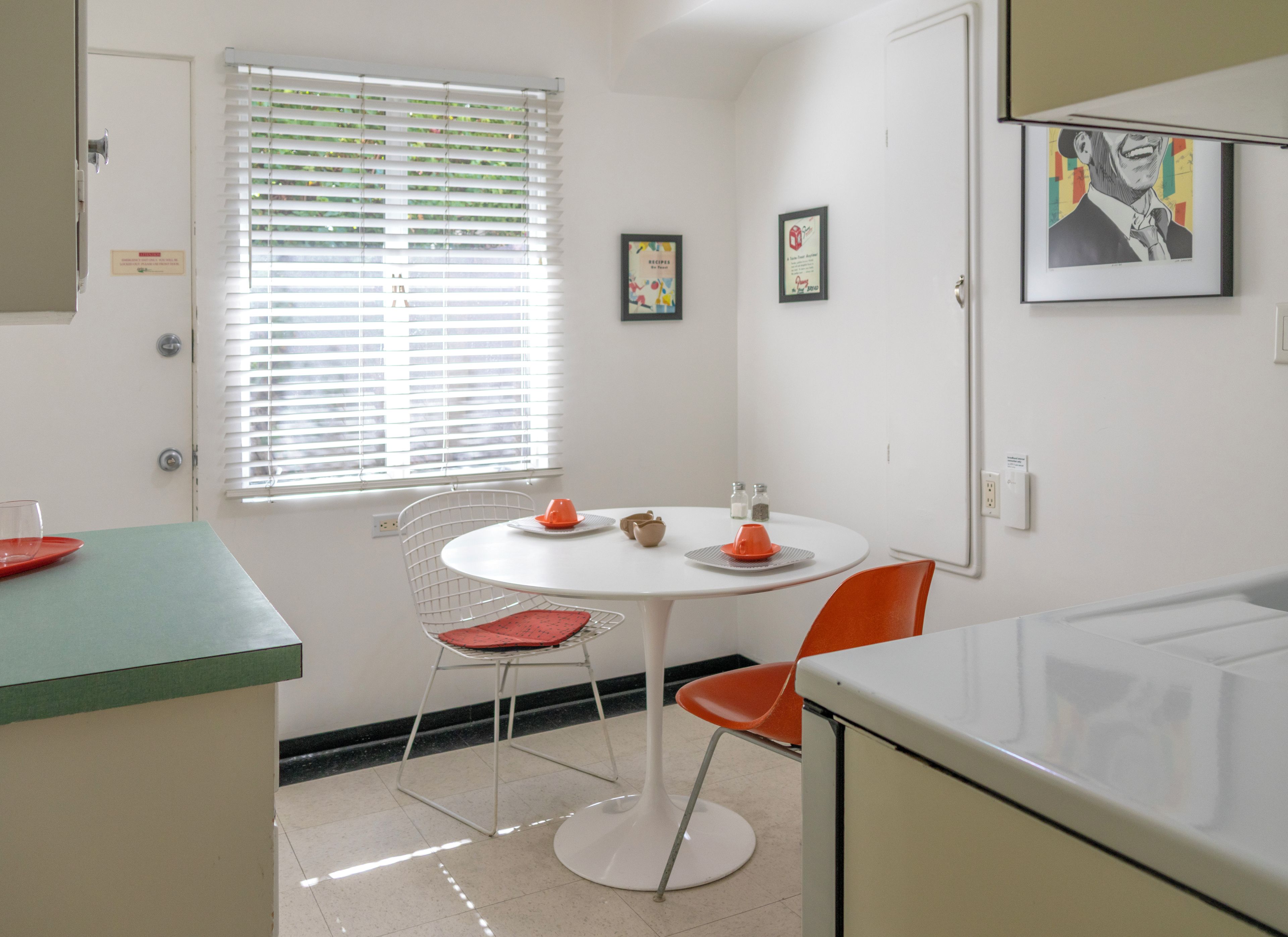 A compact kitchen with a round dining table, two chairs, a window with blinds, and a microwave.