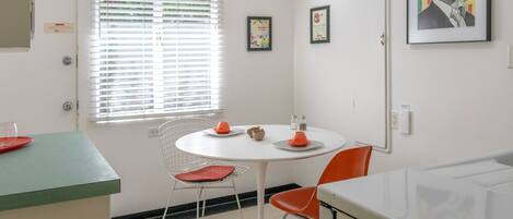 A compact kitchen with a round dining table, two chairs, a window with blinds, and a microwave.