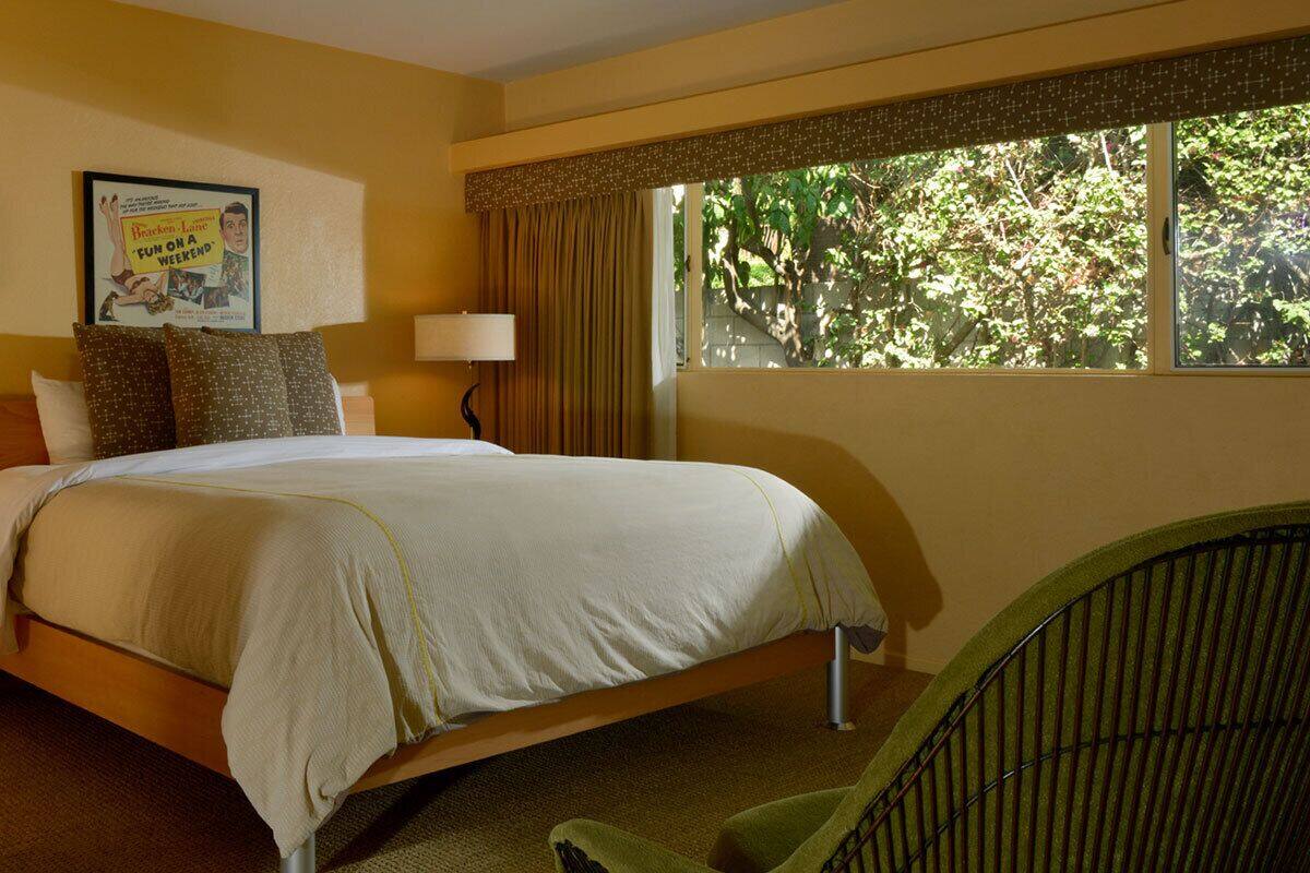 A hotel room with a bed, bedside lamps, a nightstand, a chair, and a window with a view of trees.