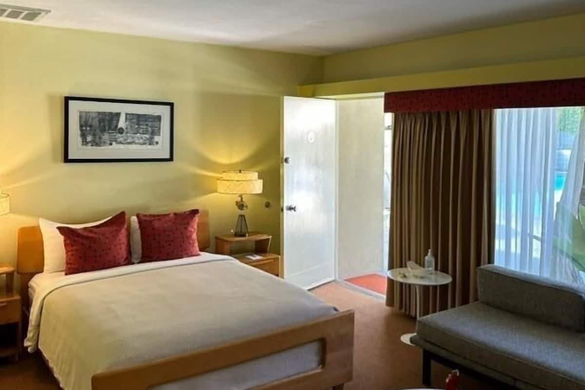 A hotel room with a bed, bedside table, lamp, and a small round table with a red vase.
