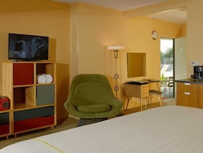 Una habitación de hotel con cama, televisor en un estante de madera, una silla verde, un escritorio con espejo y una kitchenette.