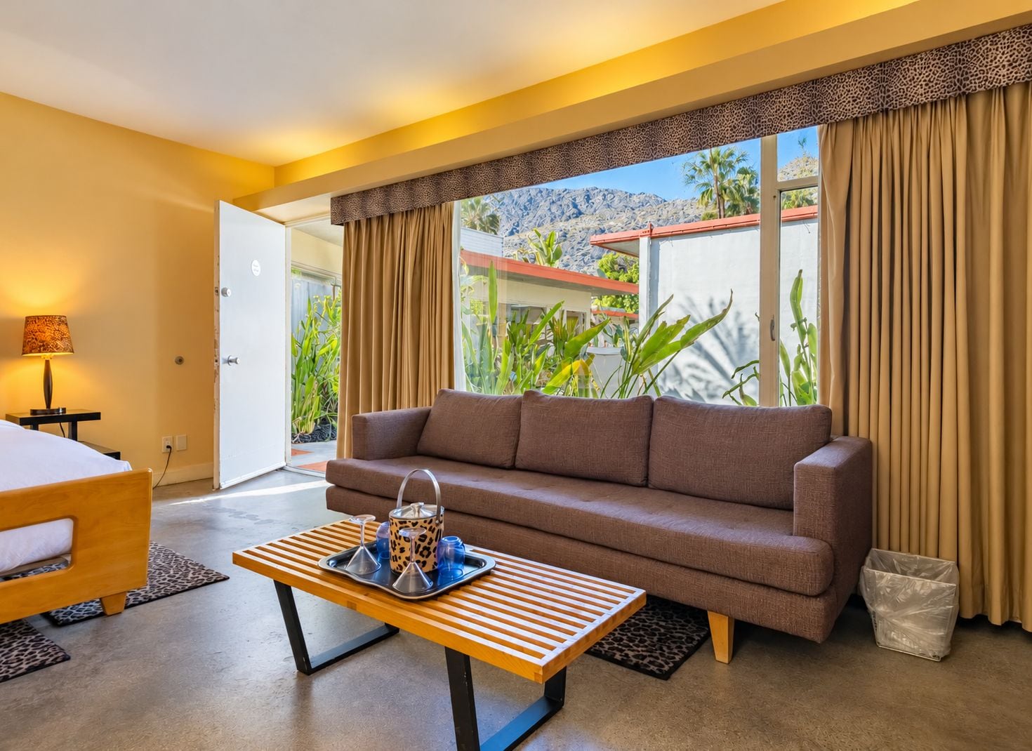 A hotel room with a sofa, a wooden coffee table with a tray, a lamp, and a view of greenery and mountains.