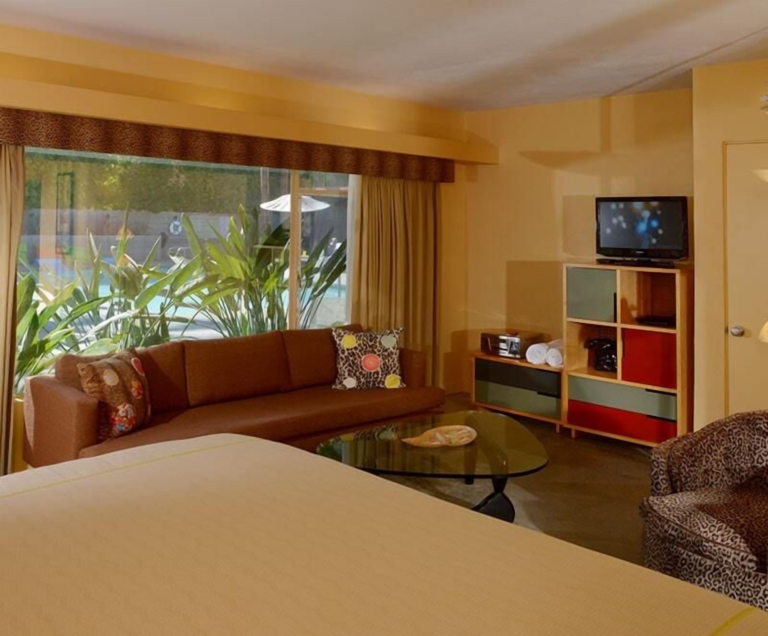 A hotel room with a sofa, a glass coffee table, a television on a stand, and a view of the outdoors through large windows.