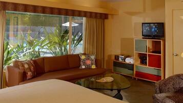 A hotel room with a sofa, a glass coffee table, a television on a stand, and a view of the outdoors through large windows.