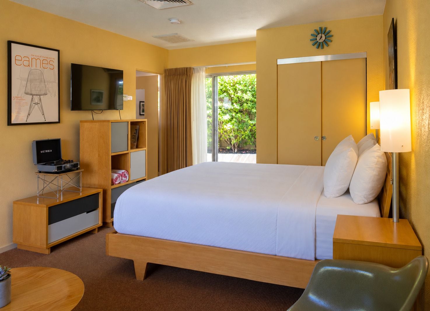 A hotel room with a large bed, a television, a desk with a computer, a bookshelf, and a window with curtains.