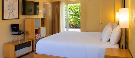 A hotel room with a large bed, a television, a desk with a computer, a bookshelf, and a window with curtains.