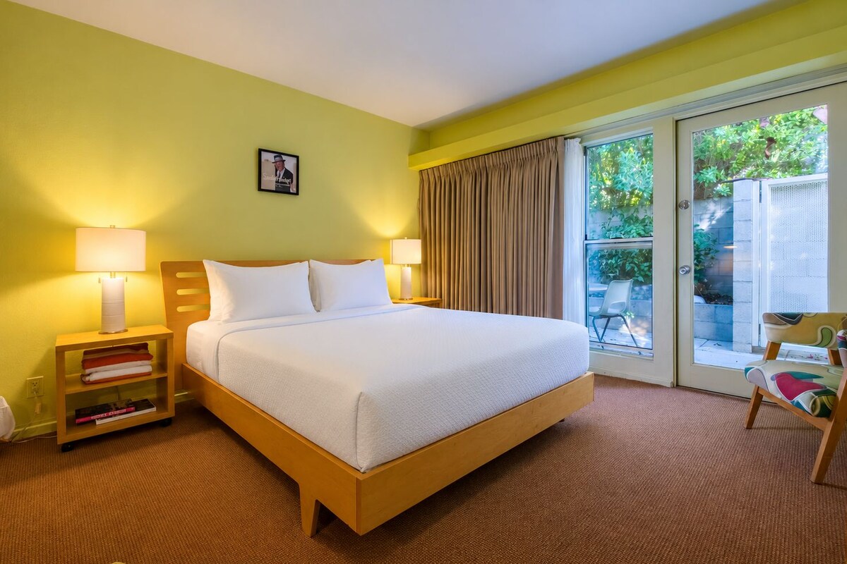 Habitación de hotel con una cama, dos lámparas de noche, una estantería, una silla y un ventanal corredizo que da a un espacio exterior.
