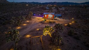 A remote desert house with a small patio, surrounded by sparse vegetation and a dirt road.