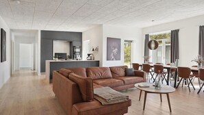 A spacious living area with a brown leather sofa, a dining table with chairs, a kitchen in the background, and a large window with curtains. - Stunning home in Blokhus with kitchen (Blokhus)