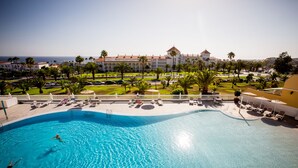 Ein großer Swimmingpool mit klarem, blauem Wasser, umgeben von Liegestühlen und Sonnenschirmen, mit Blick auf ein Hotel und Palmen im Hintergrund.