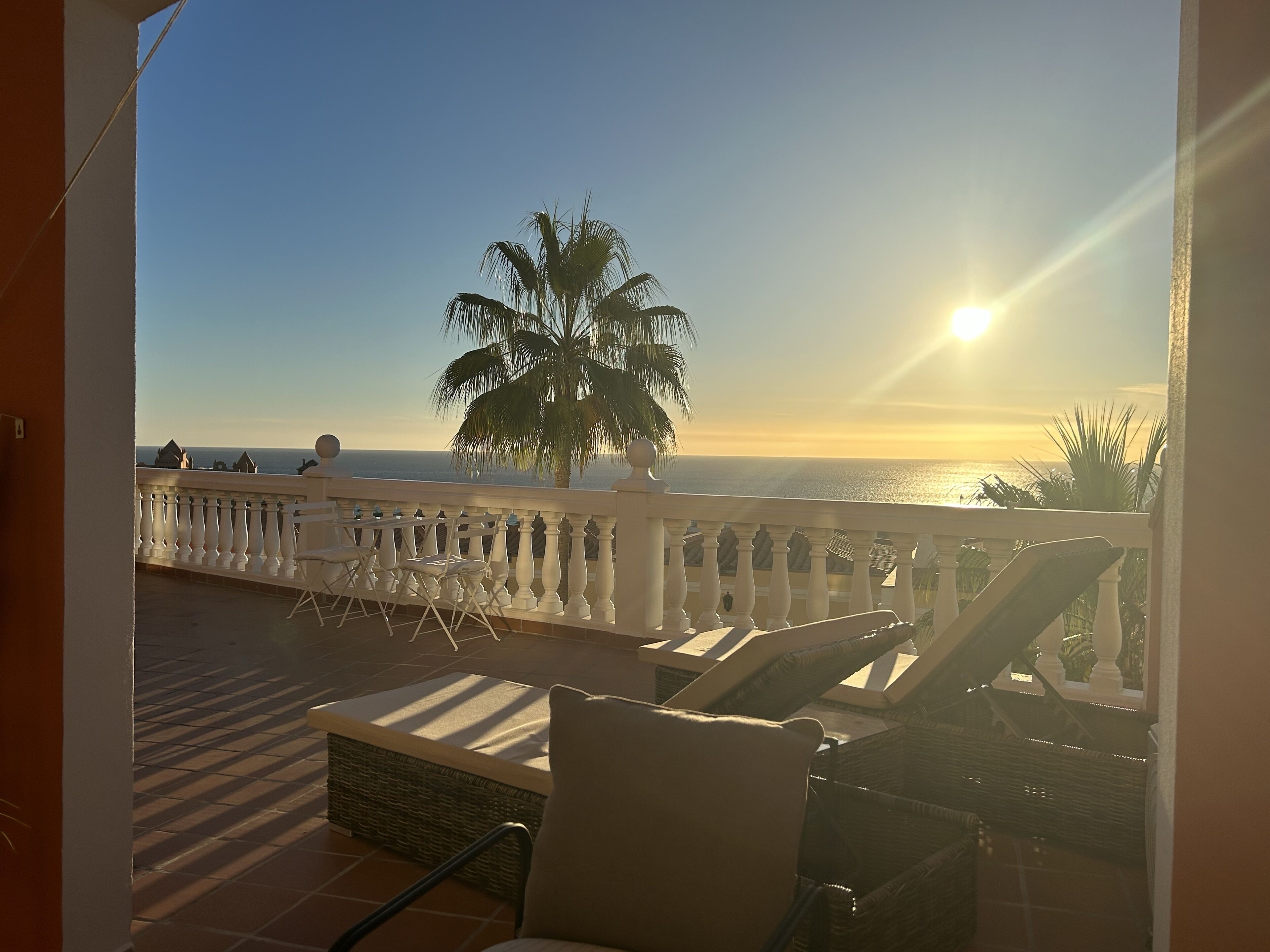 A balcony with white chairs and a wicker sofa, overlooking a sunset over the ocean.