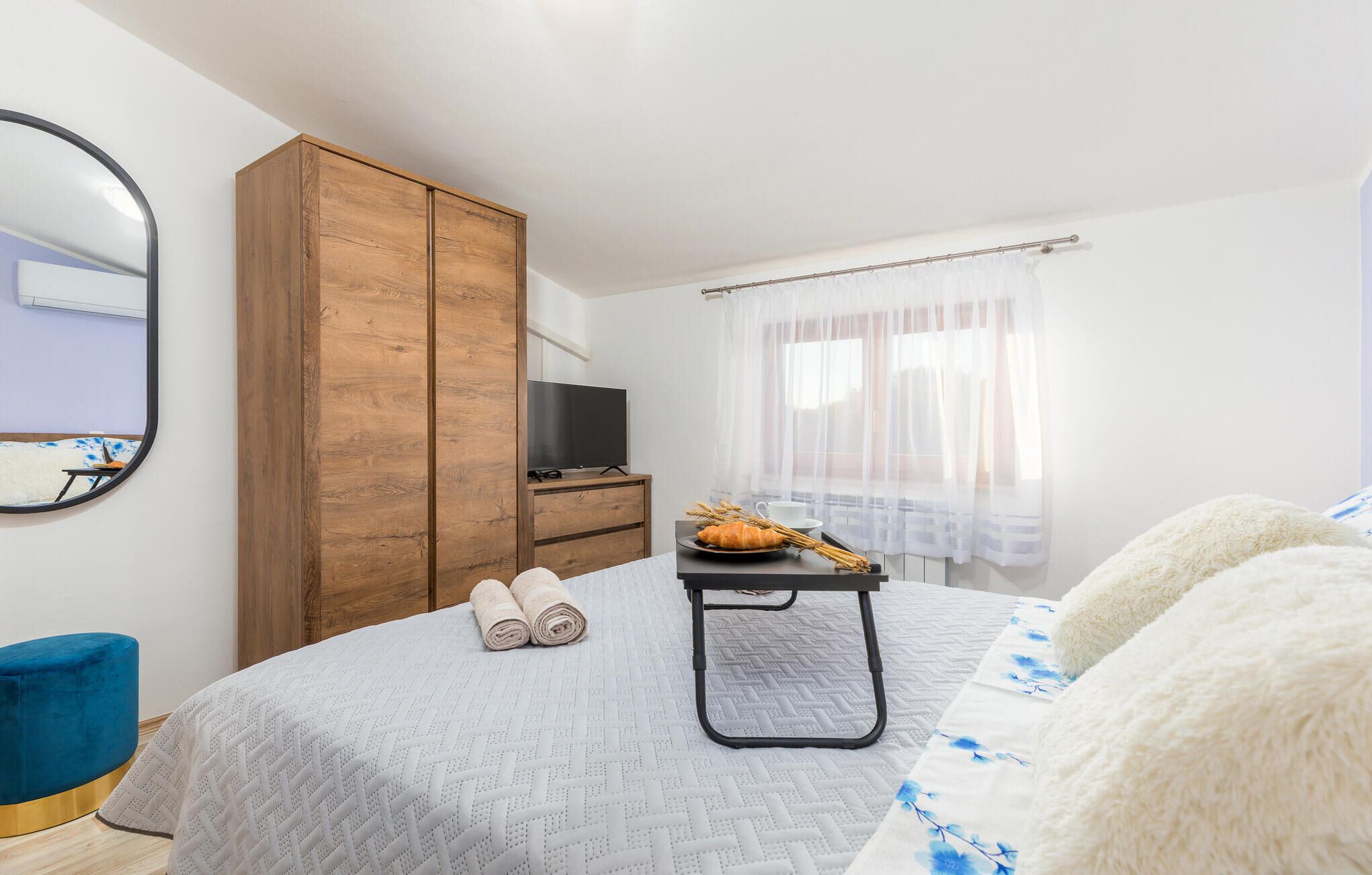 A bedroom with a bed, a wooden wardrobe, a television, a small table with a croissant and a cup, and a window with sheer curtains.